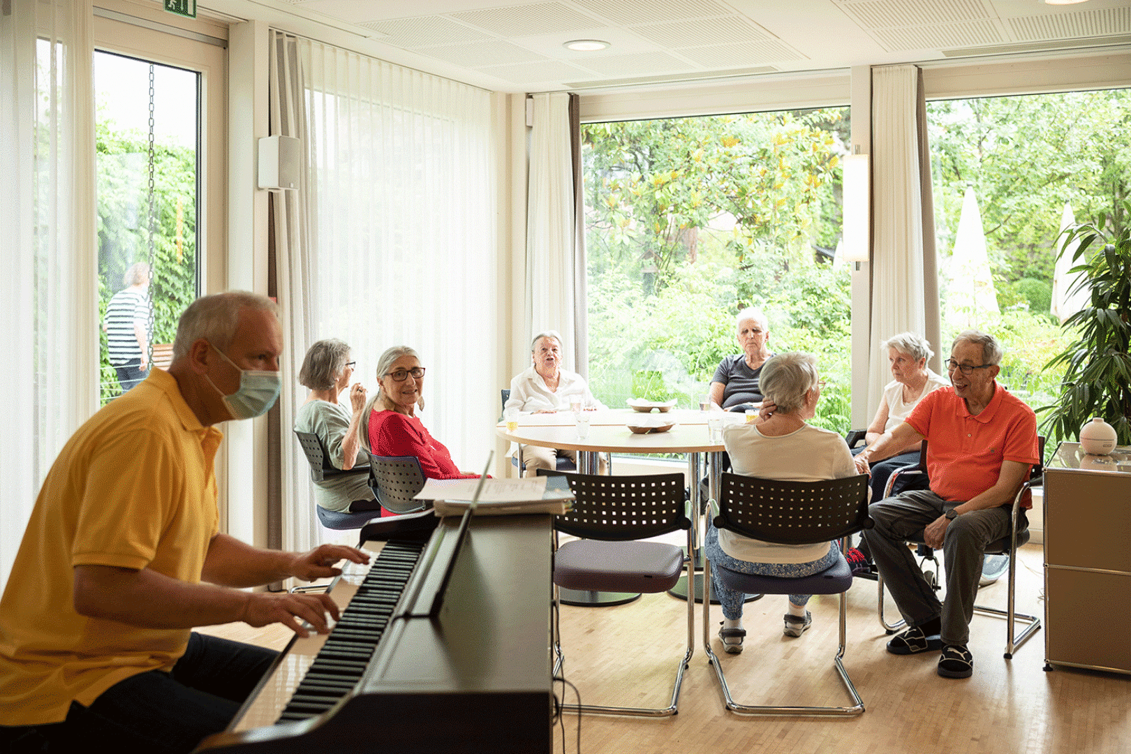 Ältere Personen sitzen an einem runden Tisch. Im Vordergrund spielt ein Mann auf einem Klavier.